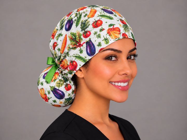 Three-quarter angle of a veggie print nurse cap, with purple eggplants and green pea pods across the gathered crown on white cotton