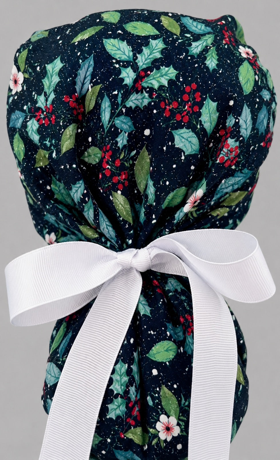 Winter holly ponytail scrub cap back view close-up showing gathered pouch full of glittery holly, red berries and blue foliage on snow-flecked navy with white ribbon bow