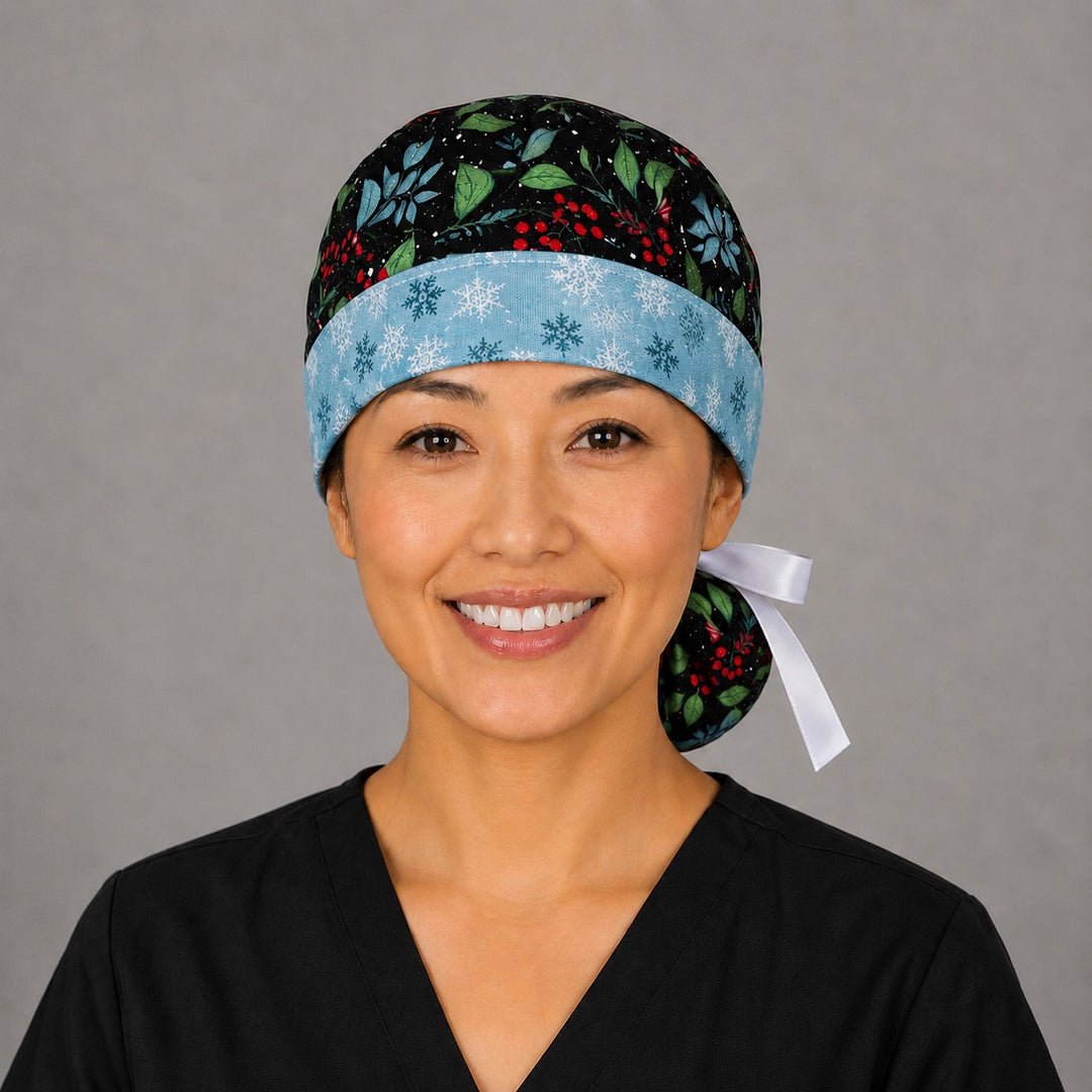 Woman wearing winter holly ponytail scrub cap, front view smiling with glittery holly, red berries and blue foliage on navy with snowflake band and white ribbon tie