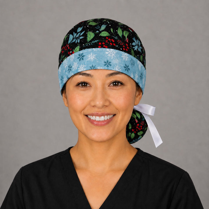 Woman wearing winter holly ponytail scrub cap, front view smiling with glittery holly, red berries and blue foliage on navy with snowflake band and white ribbon tie