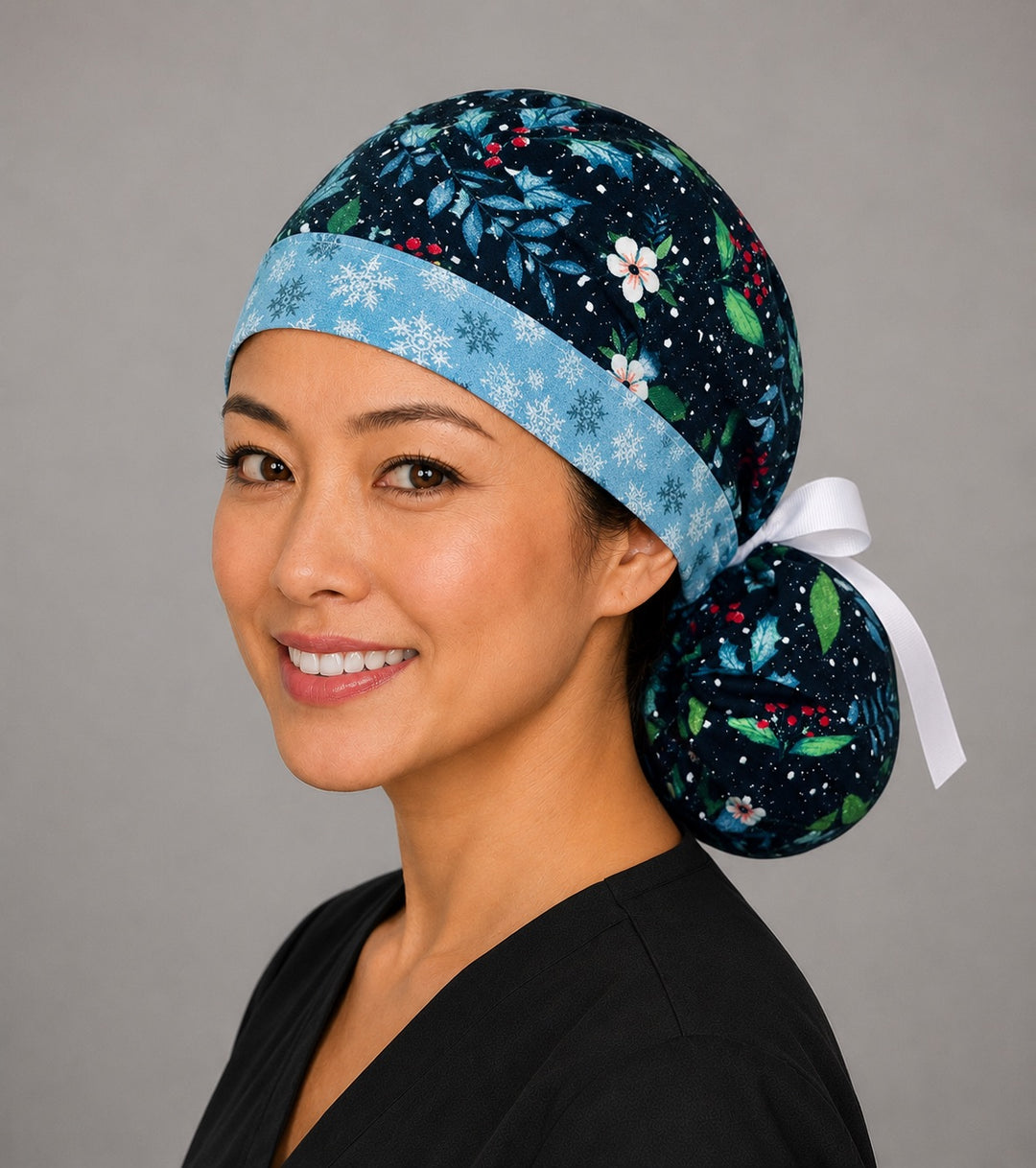 Winter holly ponytail nurse cap left three-quarter angle on smiling model showing glittery holly, red berries and blue foliage with snowflake band and white ribbon bow