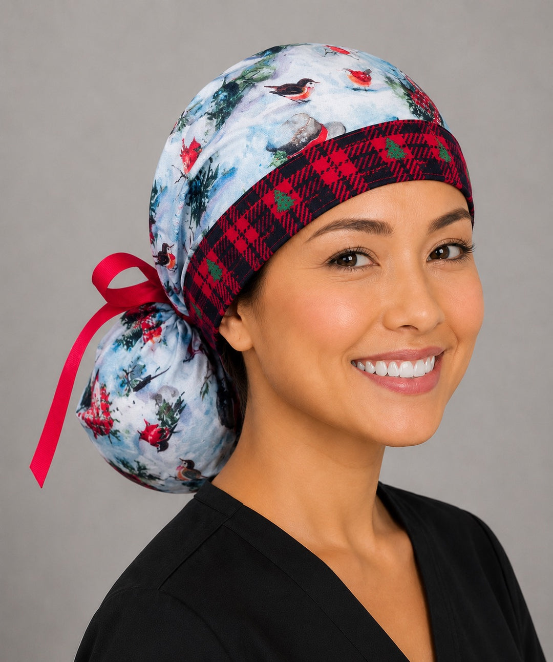 Woman wearing winter snowman ponytail scrub cap, front view smiling with snowmen, red cardinals and trees on snowy blue with red plaid band and red ribbon tie
